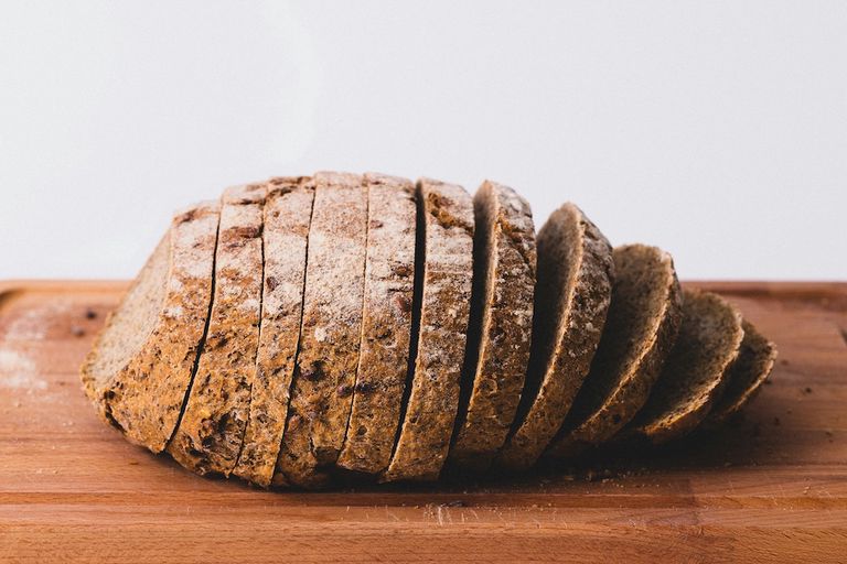 A partially sliced loaf of whole grain bread rests on a wooden cutting board, set against a plain white background.
