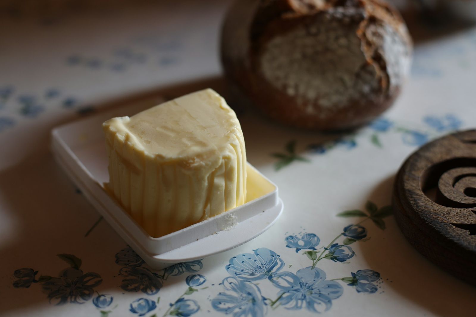 A hand-cut block of pale yellow butter on a small white dish, with a round crusty loaf of bread resting on a floral-printed cloth behind it.