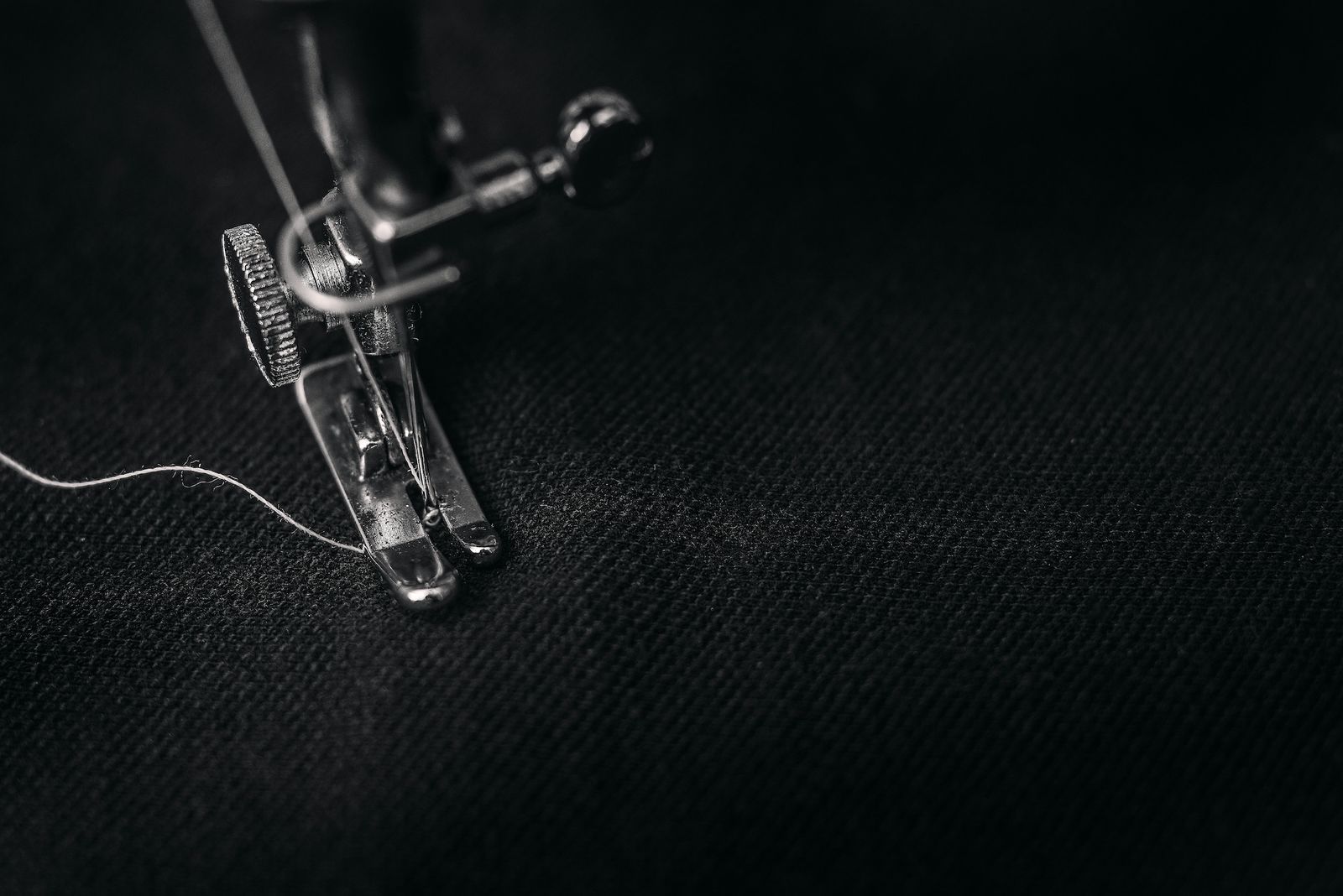 Close-up of a sewing machine needle and presser foot stitching white thread into dark fabric, showing mechanical detail and cloth texture.