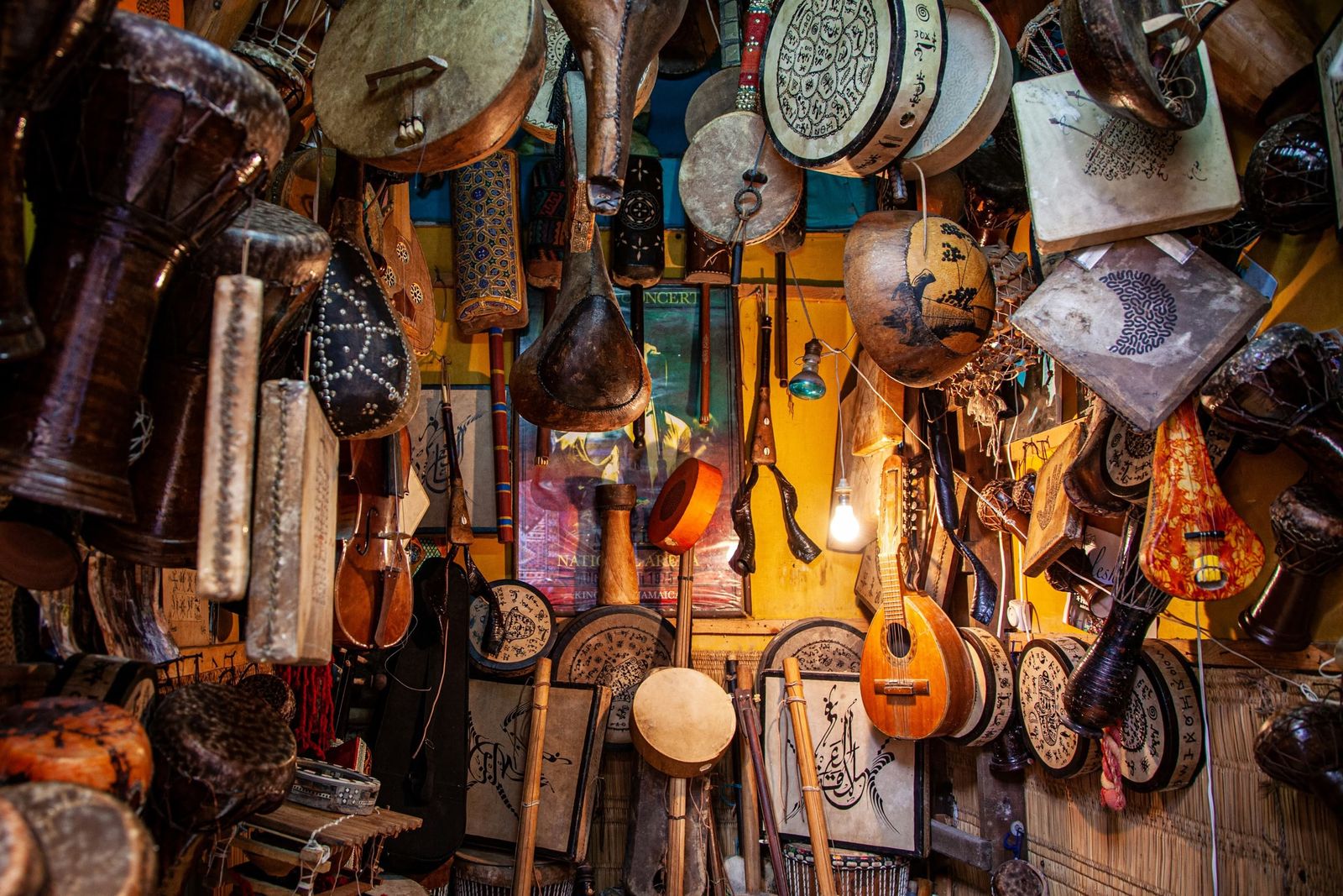 A warmly lit shop wall crowded with hanging musical instruments, including frame drums, hand drums, a mandolin, a fiddle, and painted gourds.