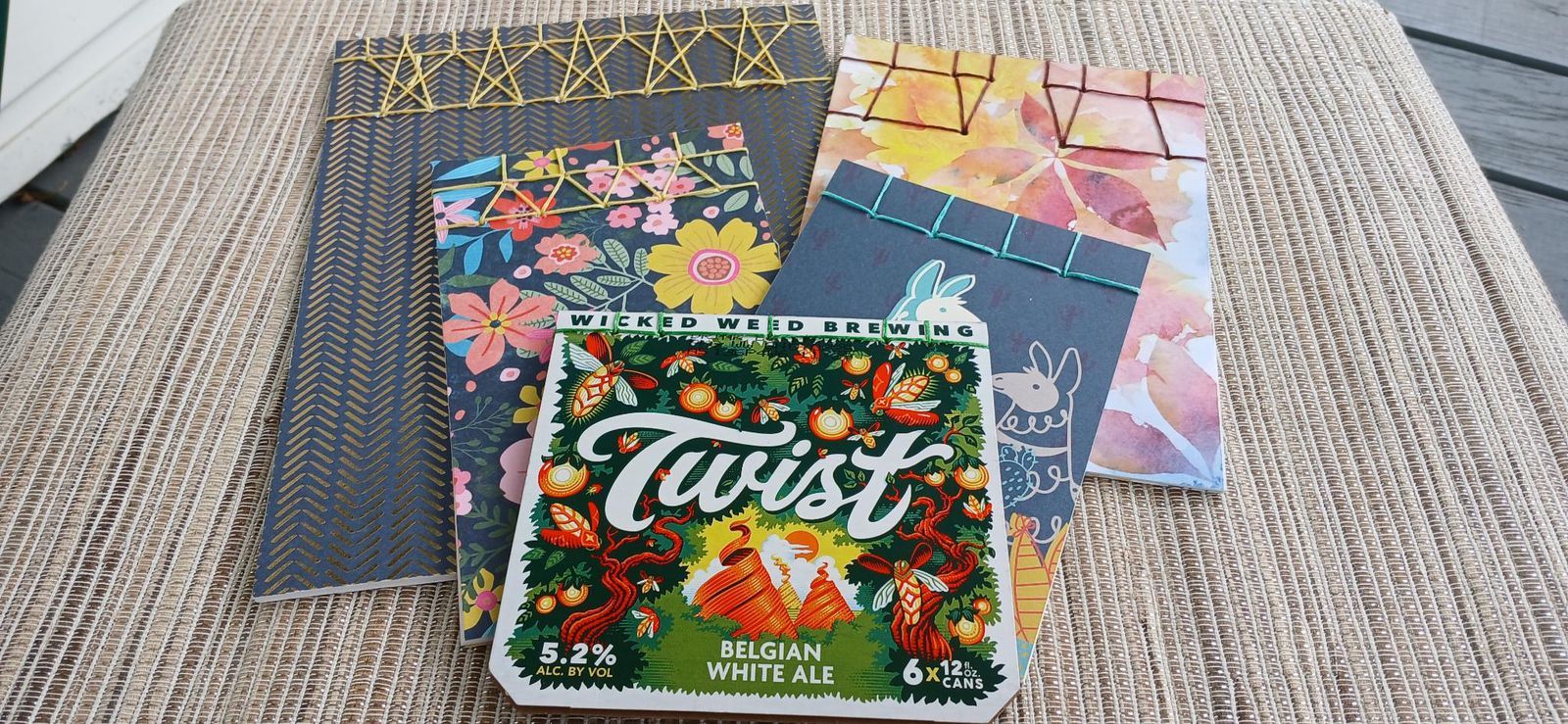 Several handmade journals with colorful patterned covers arranged on a woven mat, each bound along the top edge with a decorative Japanese stab-binding stitch in gold, green, and blue thread.
