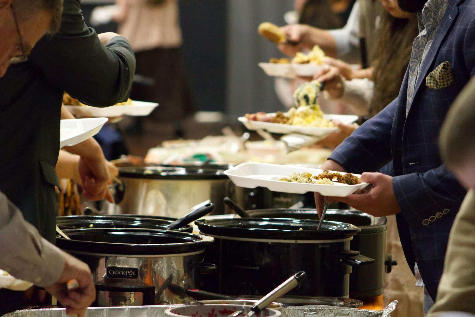 People moving down a potluck line, filling their plates from a row of slow cookers and covered dishes.