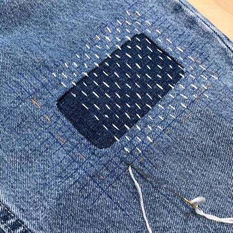 A pair of jeans with a denim patch hand-sewn on using rows of white sashiko running stitches.