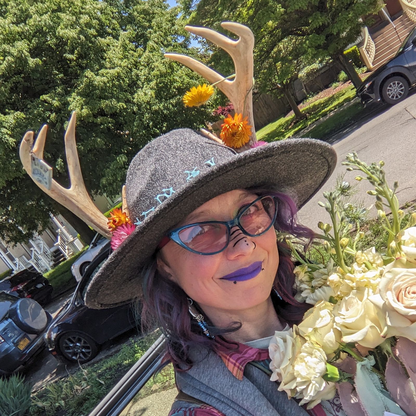 A person with purple lipstick and glasses wears a wide hat with antlers, flowers, embroidery; holds white roses outside on a sunny day.