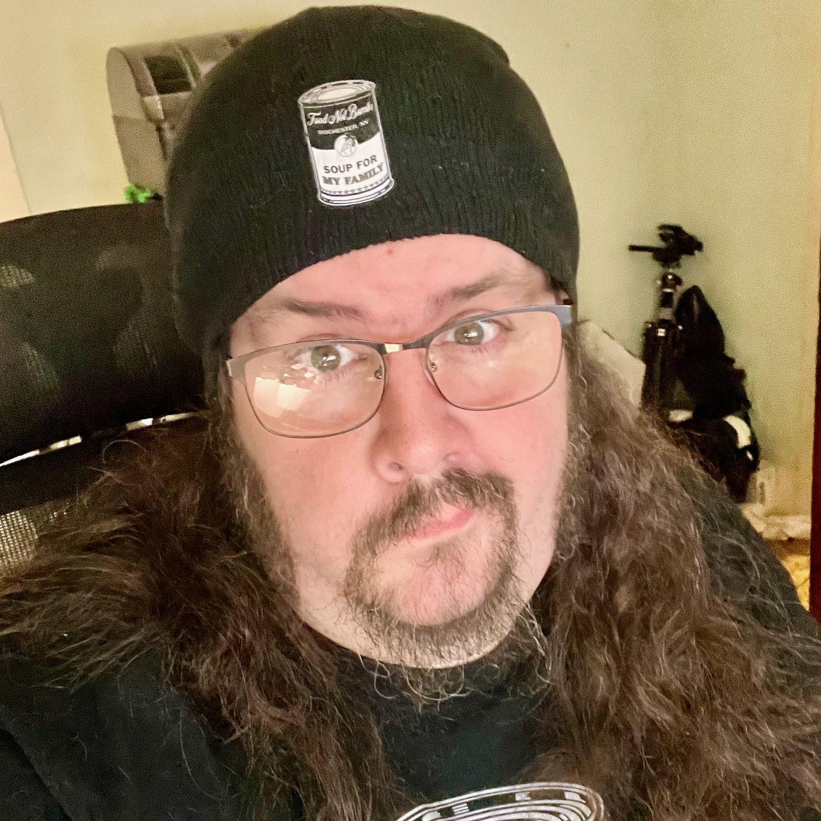 A person with long brown hair, glasses, and a black beanie with a soup can on it that reads 'Soup for My Family' sits in an office chair reading. A tripod and 3D printer sit in the background.