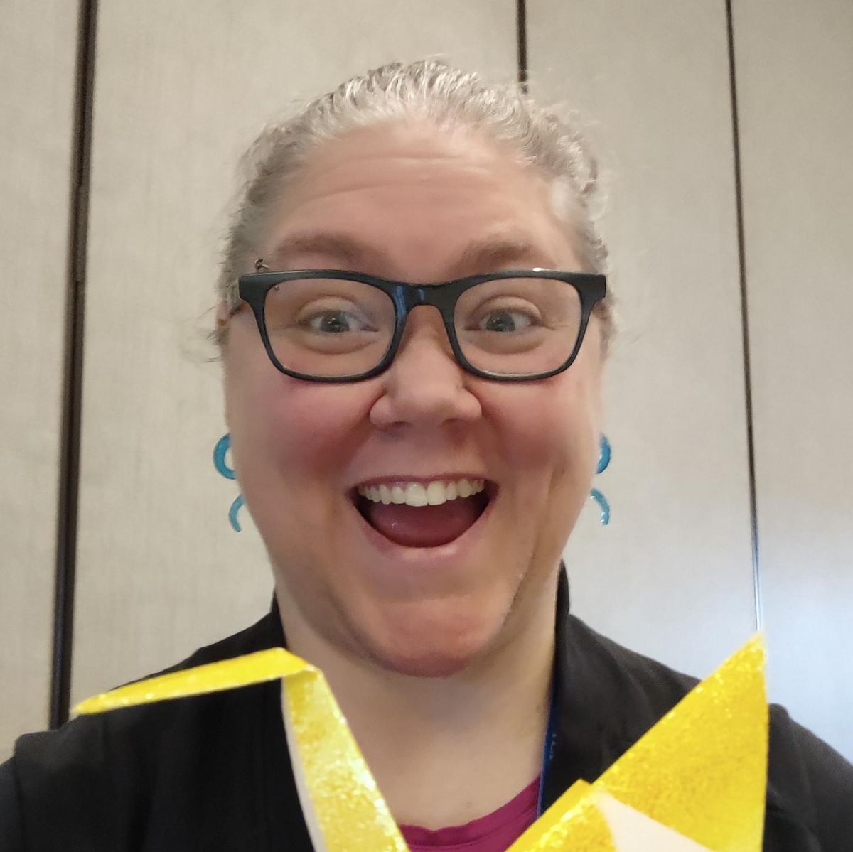 A smiling person with glasses and blue earrings holds a large yellow origami crane indoors, near a beige panel wall.