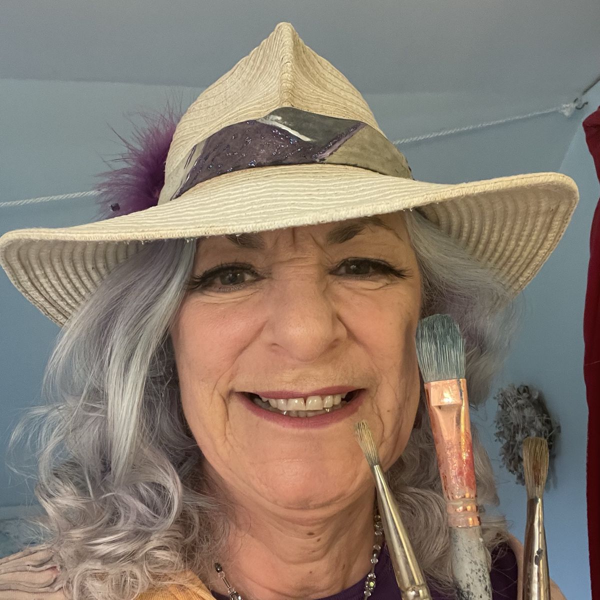 Smiling woman with long gray hair and a wide smile, wearing a wide-brimmed hat with a purple band, holds paintbrushes in front of her face.