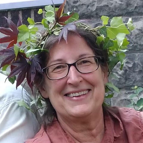 A woman with glasses smiles, wearing a crown of green and red leaves. She stands in front of a stone wall.