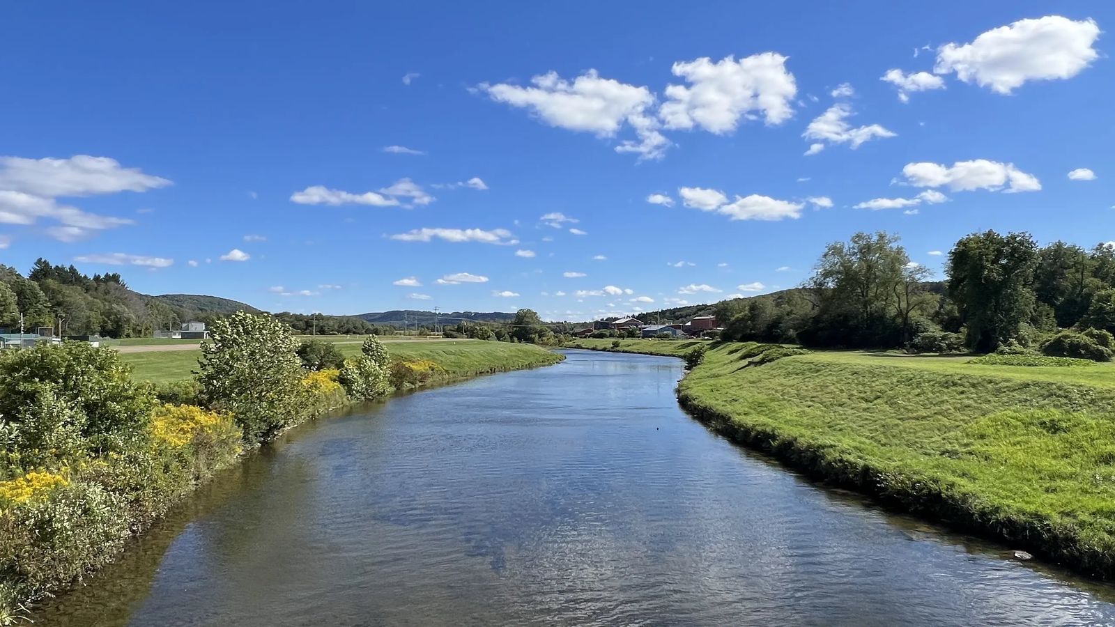 View of a calm river running between green grassy banks under a partly cloudy sky in Allegany County, NY.